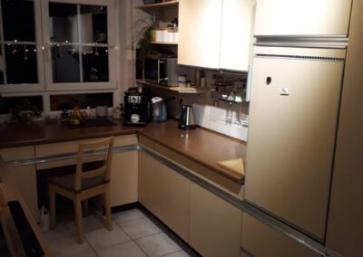 Eine gemütliche Küche mit beigen Schränken, einer braunen Arbeitsplatte, einem Holzstuhl am Fenster und verschiedenen Geräten wie Kaffeemaschine und Wasserkocher. Vor dem Fenster sind Lichterketten zu sehen, und die Küche ist warm beleuchtet.