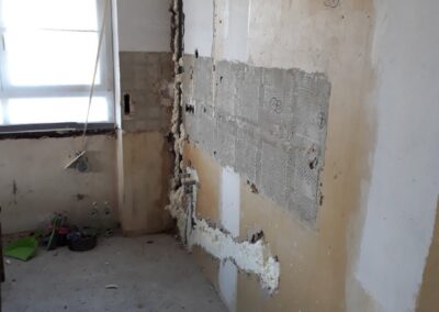 Ein teilweise abgerissener Raum mit freiliegendem Putz und Isolierung an den Wänden, fehlenden Fliesen, Bauschutt auf dem Boden und einem Fenster, das natürliches Licht hereinlässt.