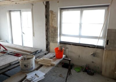 Ein Raum im Umbau mit freiliegenden Wänden, Arbeitstischen, Werkzeugen, Eimern, Holzbrettern, Papieren und verstreuten Baumaterialien in der Nähe großer Fenster, die Tageslicht hereinlassen.
