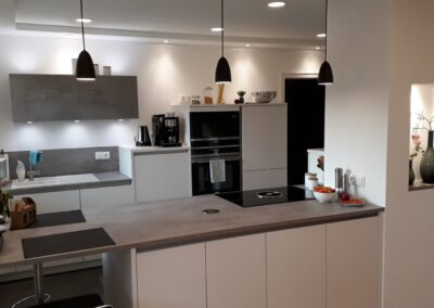 Moderne Küche mit weißen Schränken, Einbaugeräten, grauen Arbeitsplatten und Pendelleuchten. Auf den Arbeitsflächen und Regalen stehen Pflanzen, eine Kaffeemaschine und Schüsseln. In einer Wandnische steht eine dekorative Vase.