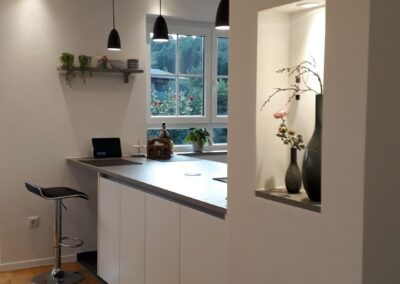 Moderne Küche mit hellem Holzboden, weißen Schränken, schwarzen Pendelleuchten, einem Barhocker und Pflanzen auf der Arbeitsplatte. Ein Fenster sorgt für Tageslicht; an der Wand ist eine dekorative Nische mit Vasen und einem Zweig sichtbar.