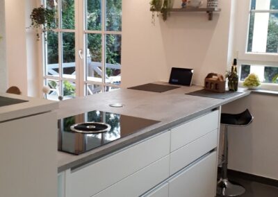 Moderne Küche mit weißen Schränken, einer grauen Kücheninsel, Barhocker, Hängelampen und einem Fenster mit Blick ins Grüne. Pflanzen und ein kleines Regal mit Gegenständen schmücken den Raum und schaffen eine gemütliche Atmosphäre.