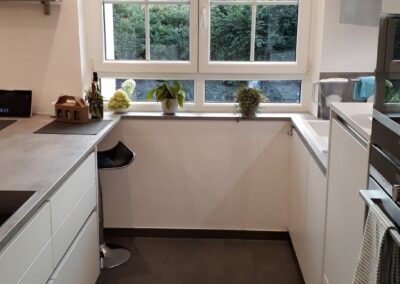 Moderne Küche mit weißen Schränken, grauen Arbeitsplatten, einem gemusterten Teppich und einem großen Fenster mit Blick auf grünes Laub. Auf der Fensterbank stehen Pflanzen, an der Theke steht ein schwarzer Hocker.