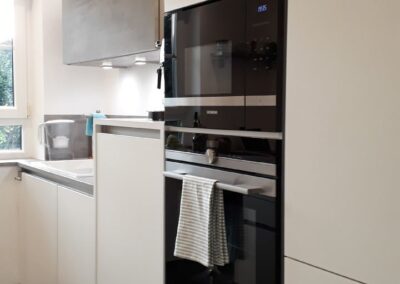 Moderne Küche mit weißen Schränken, eingebautem Backofen und Mikrowelle, einem Handtuch am Backofengriff und Gläsern mit Nudeln und Getreide auf den Schränken. Tageslicht fällt durch ein Fenster links ein.