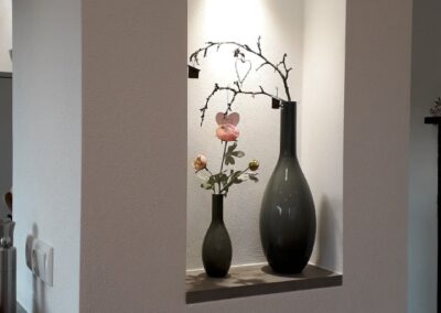In einer vertieften Wandnische sind zwei Vasen ausgestellt: eine hohe dunkle Vase mit einem Zweig mit kleinen Blüten und eine kleinere Vase mit einer einzelnen rosa Blüte. Beide werden von einem kleinen Scheinwerfer darüber beleuchtet.