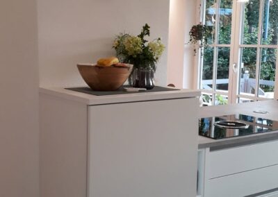 Moderne Küche mit weißen Schränken, einer Holzschale mit Zitronen und einer Glasvase mit Blumen auf der Arbeitsplatte. An der Wand darüber hängt ein dekorativer Tierkopf. Große Fenster lassen Tageslicht herein.